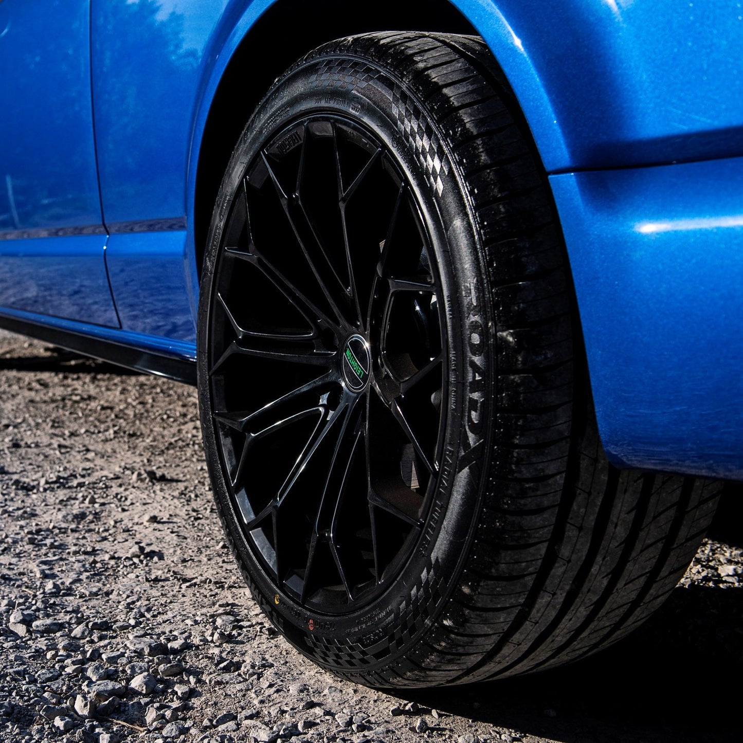 Close-up of a sleek black alloy wheel on a blue car, showcasing a modern design on a gravel surface.