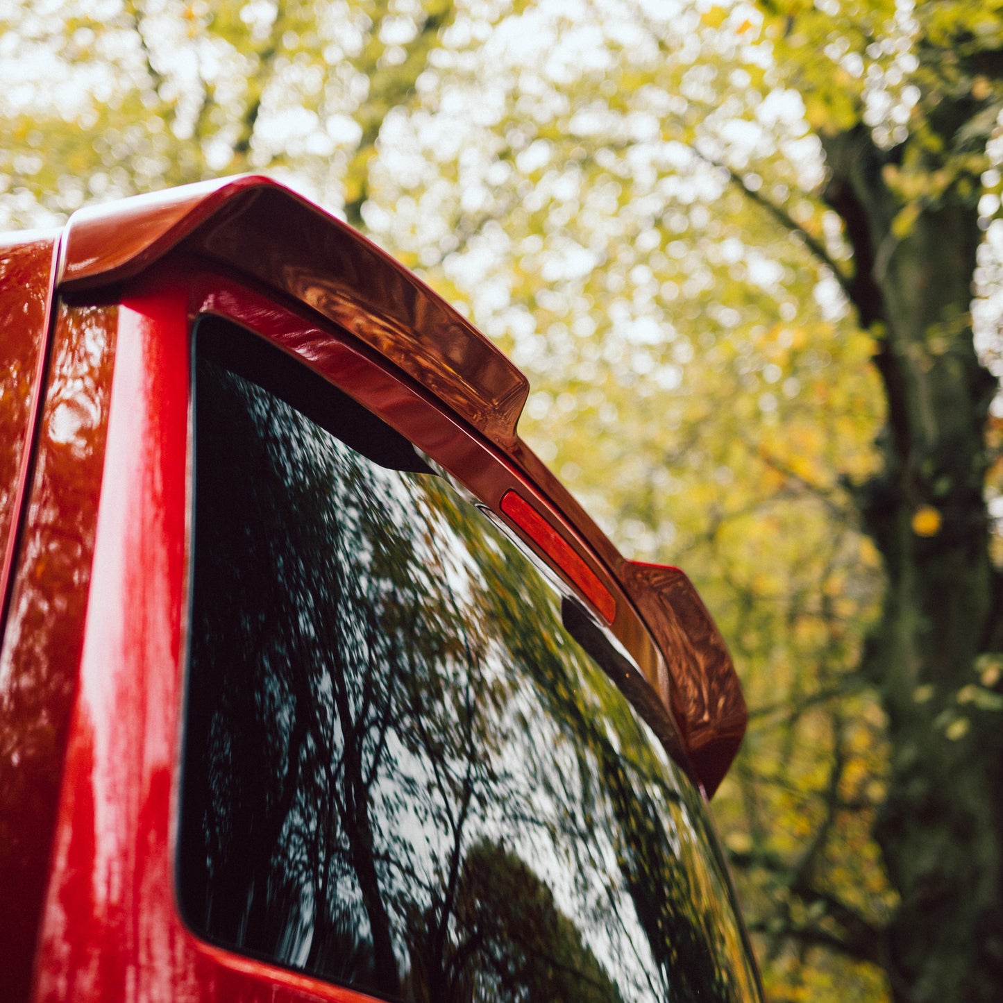 Close-up of a red vehicle's rear with a sporty spoiler and reflections of trees in the tinted glass, set against a vibrant autumn backdrop.