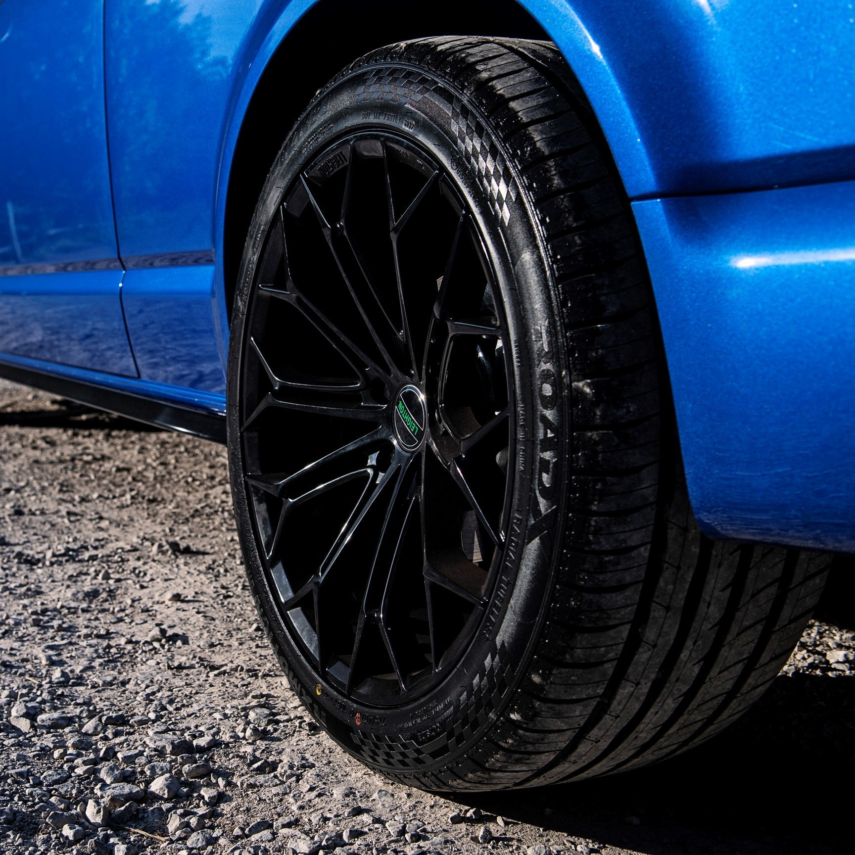 Close-up of a sleek black alloy wheel on a blue car, showcasing a modern design on a gravel surface.