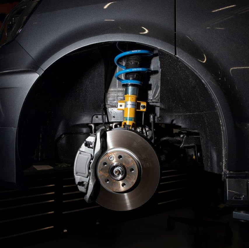 Close-up of a car's front wheel assembly with a metallic brake disc, shock absorber, and suspension components in an automotive shop.