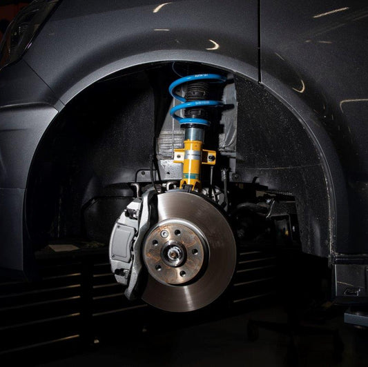 Close-up of a car's front wheel assembly with a metallic brake disc, shock absorber, and suspension components in an automotive shop.