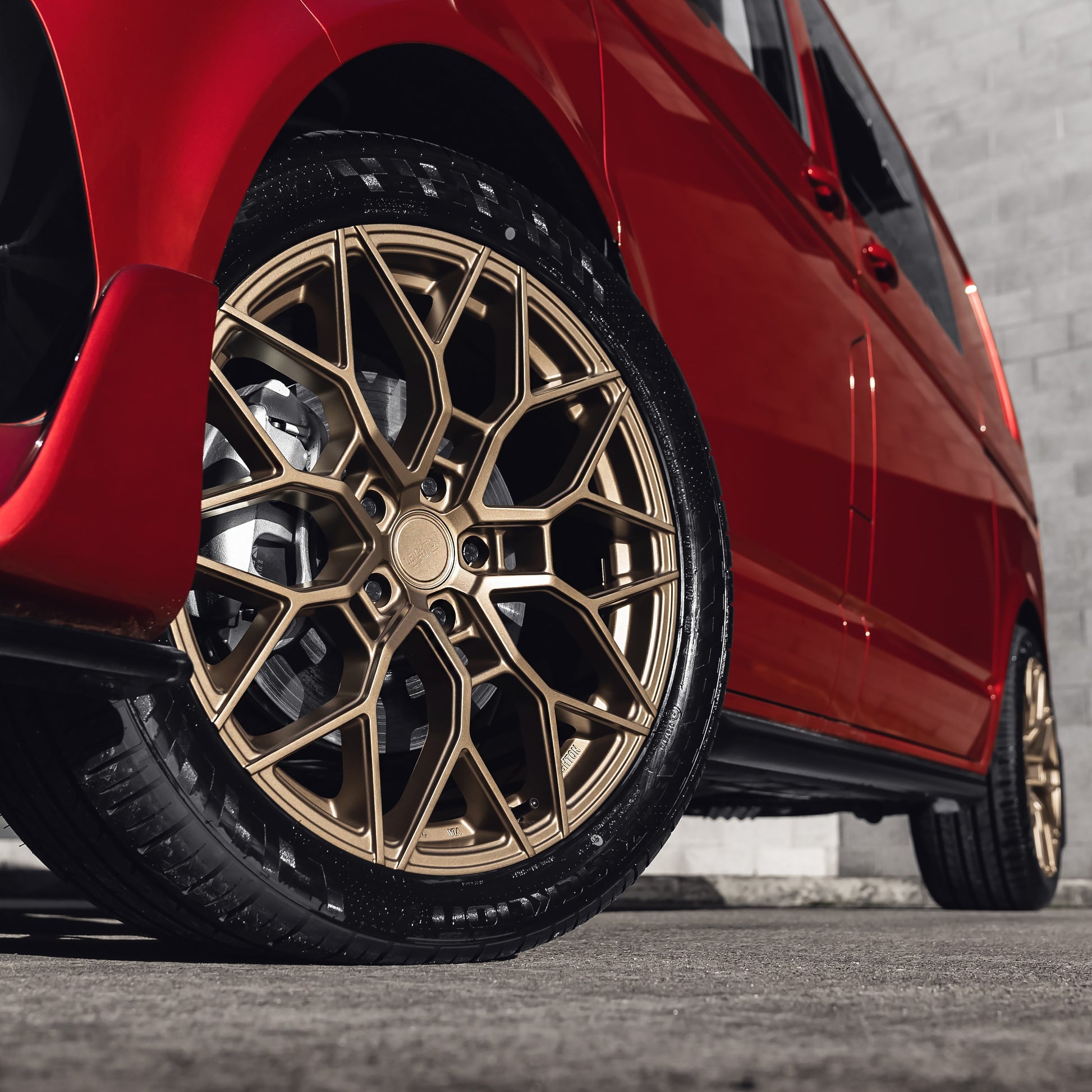 A close-up of a red car's gold alloy wheel with intricate design, parked on a smooth surface against a textured wall background.