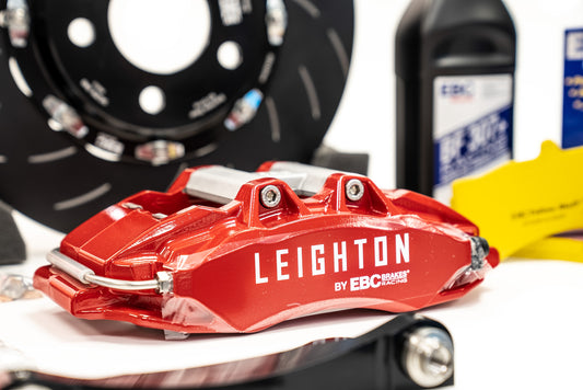 Close-up of a bright red Leighton brake caliper by EBC Racing, placed on a white surface with brake components in the background.