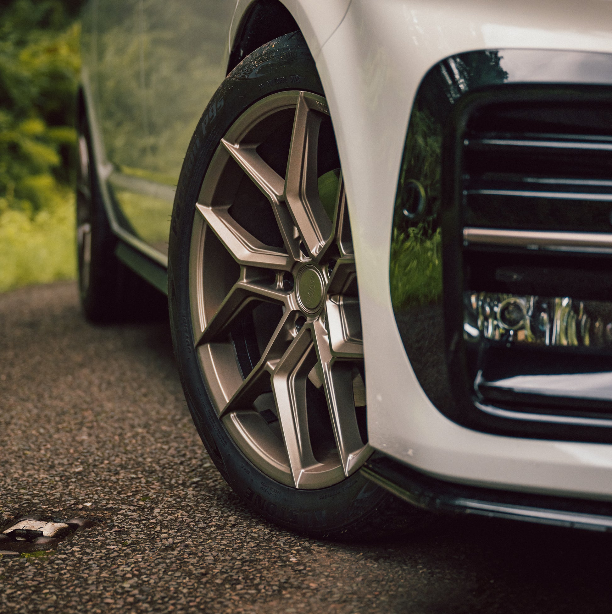 Close-up of a stylish, angular alloy wheel on a white car, parked on a gravel road with lush greenery in the background.
